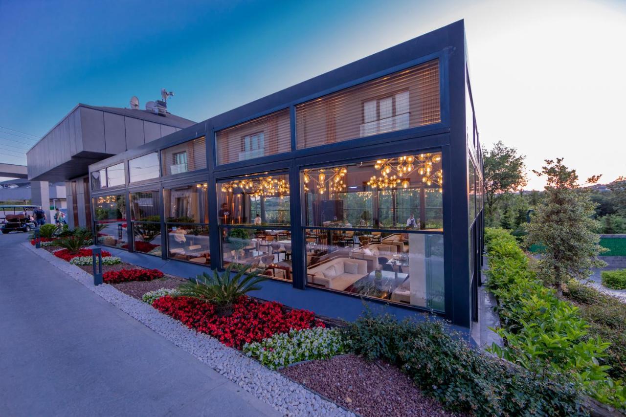 Menalo Hotel Premium Istanbul Airport Exterior photo The photo shows a modern restaurant or dining space characterized by large glass windows that create an open and inviting atmosphere. The exterior features a sleek, dark frame with a contemporary design. There are vibrant flower beds with red and gre