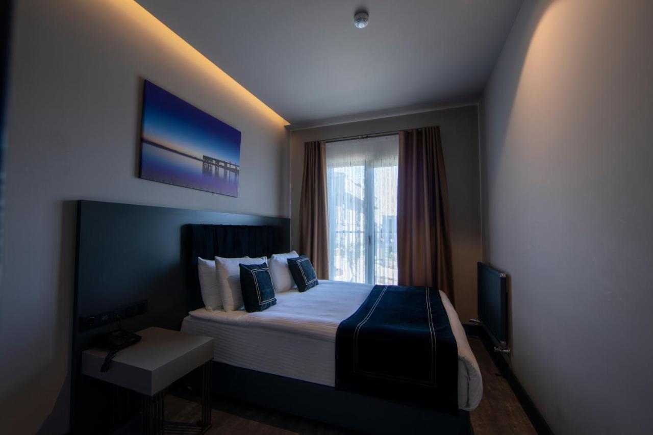 Menalo Hotel Premium Istanbul Airport Exterior photo The photo shows a modern hotel room featuring a double bed with neatly arranged pillows and bedding in white and navy blue colors. To the left, there is a small bedside table. A flat-screen TV is mounted on the wall across from the bed. Large windows