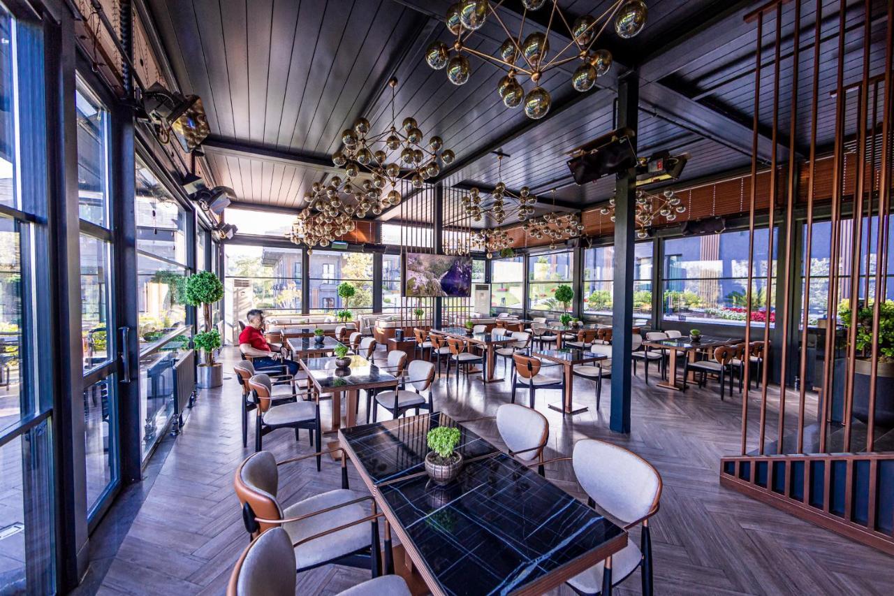 Menalo Hotel Premium Istanbul Airport Exterior photo The photo shows a stylish restaurant interior that features modern decor. There are several tables arranged around the space, each with elegant seating. The tables are set with small green plants as centerpieces. The ceiling is adorned with decorativ