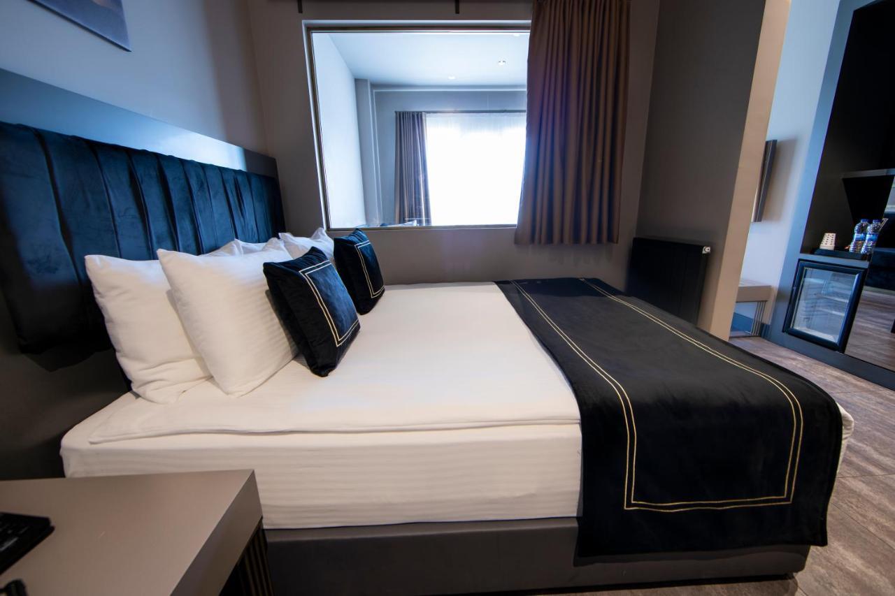 Menalo Hotel Premium Istanbul Airport Exterior photo The photo shows a modern hotel room with a neatly made bed. The bed is dressed in white linens, with a dark blanket and decorative pillows. There's a large headboard behind the bed and a bedside table on one side. Natural light is streaming in throug