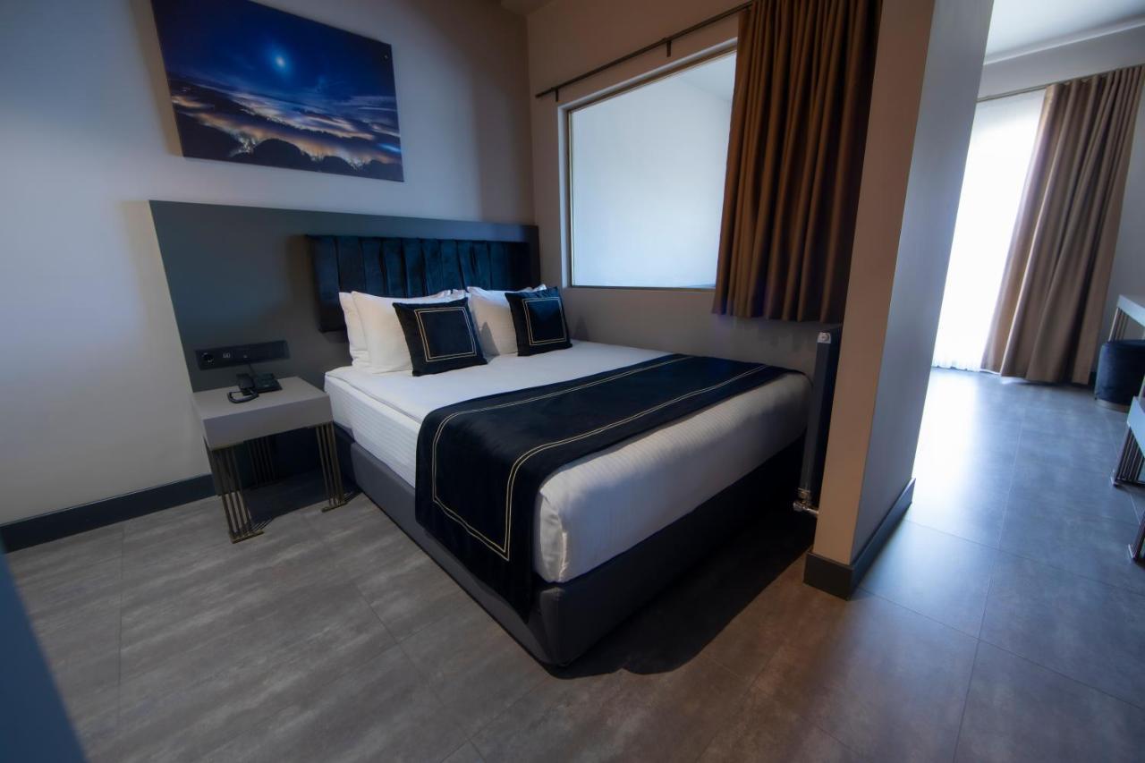 Menalo Hotel Premium Istanbul Airport Exterior photo The photo shows a modern hotel room featuring a double bed with black and white bedding. There are two decorative pillows on the bed, and behind it is a wall-mounted headboard. To the left of the bed, there's a small bedside table with a lamp. A larg